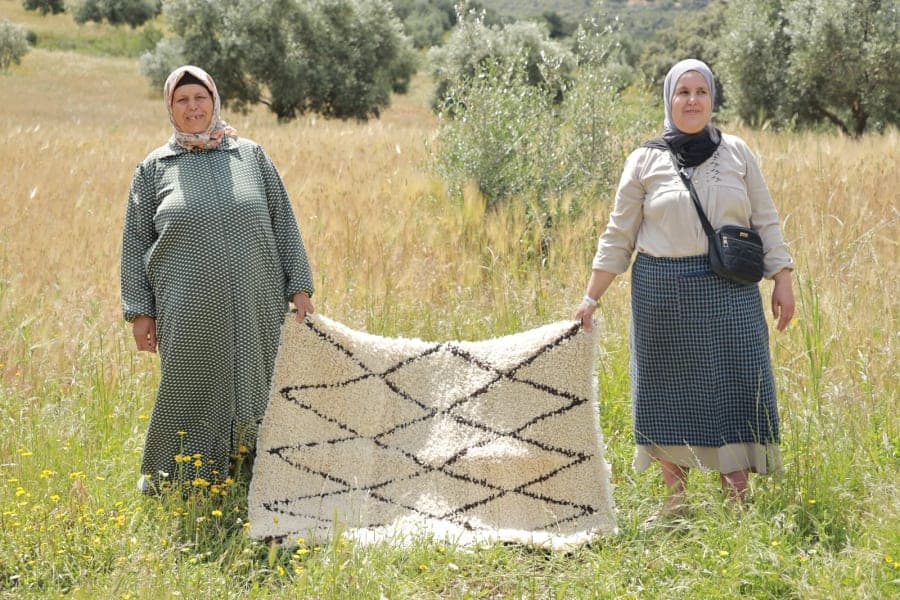 Deux femmes présentent un tapis clair à motifs sombres dans un paysage de collines.