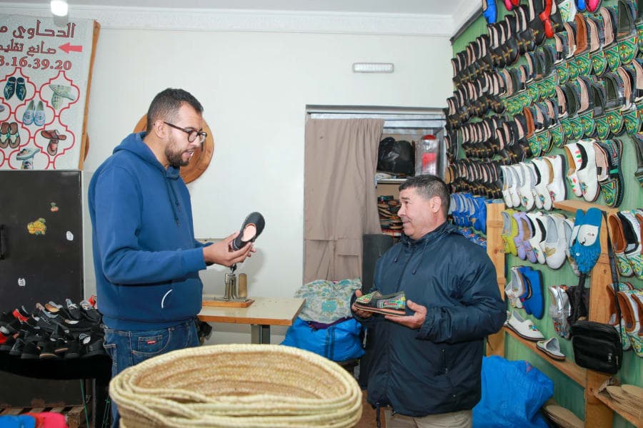 Deux hommes échangent autour d’un petit objet dans un espace de présentation de produits artisanaux.