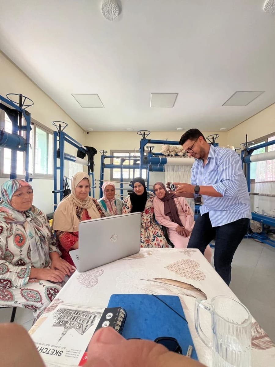 Un intervenant présente un téléphone à un groupe de femmes réunies autour d’un ordinateur entre des métiers à tisser.