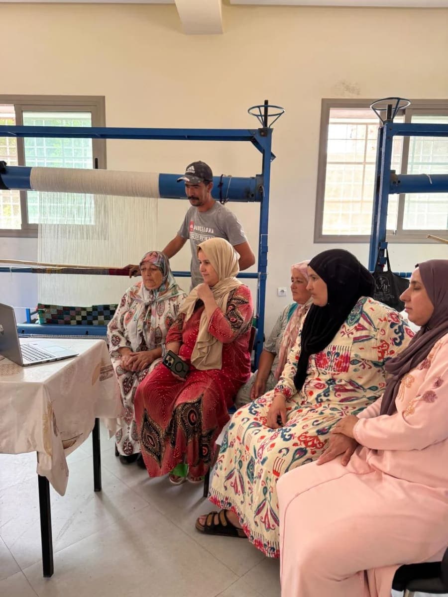 Plusieurs femmes sont réunies dans un atelier équipé de métiers à tisser bleus, avec un intervenant debout derrière elles.
