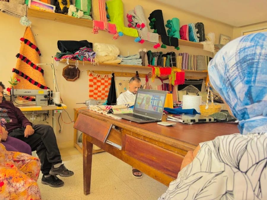 Un ordinateur affiche le portrait d’une artisane dans un atelier entouré de textiles colorés, de fils et d’une machine à coudre.