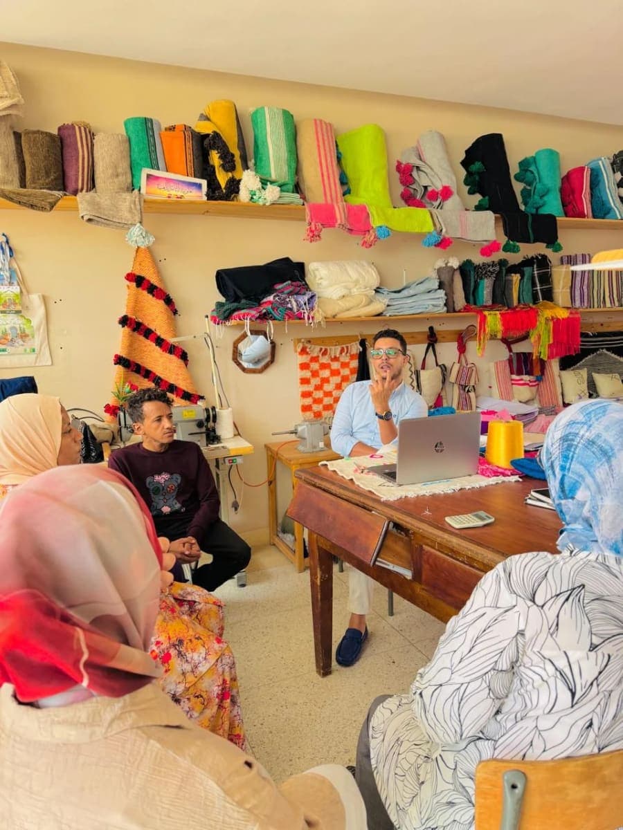 Un intervenant assis derrière un ordinateur échange avec un groupe dans un atelier rempli de créations textiles.